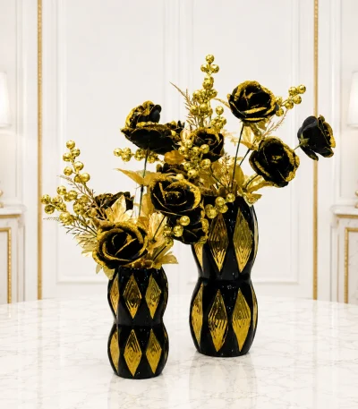 A set of two black and gold artificial floral arrangements featuring black roses with gold glitter edges and gold berry sprigs in matching hourglass-shaped black ceramic vases with gold diamond patterns, standing on a marble table