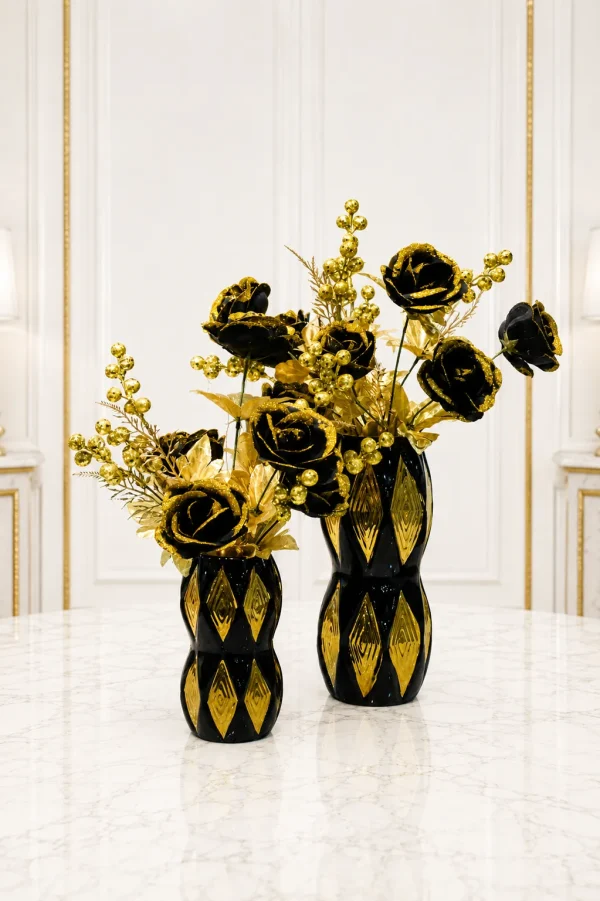 A set of two black and gold artificial floral arrangements featuring black roses with gold glitter edges and gold berry sprigs in matching hourglass-shaped black ceramic vases with gold diamond patterns, standing on a marble table