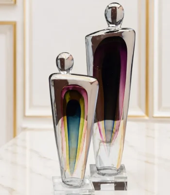 A pair of abstract art glass sculptures of differing heights, featuring clear thick glass shapes with embedded multi-colored cores of purple, blue, yellow, and pink, standing on square crystal bases on a marble table
