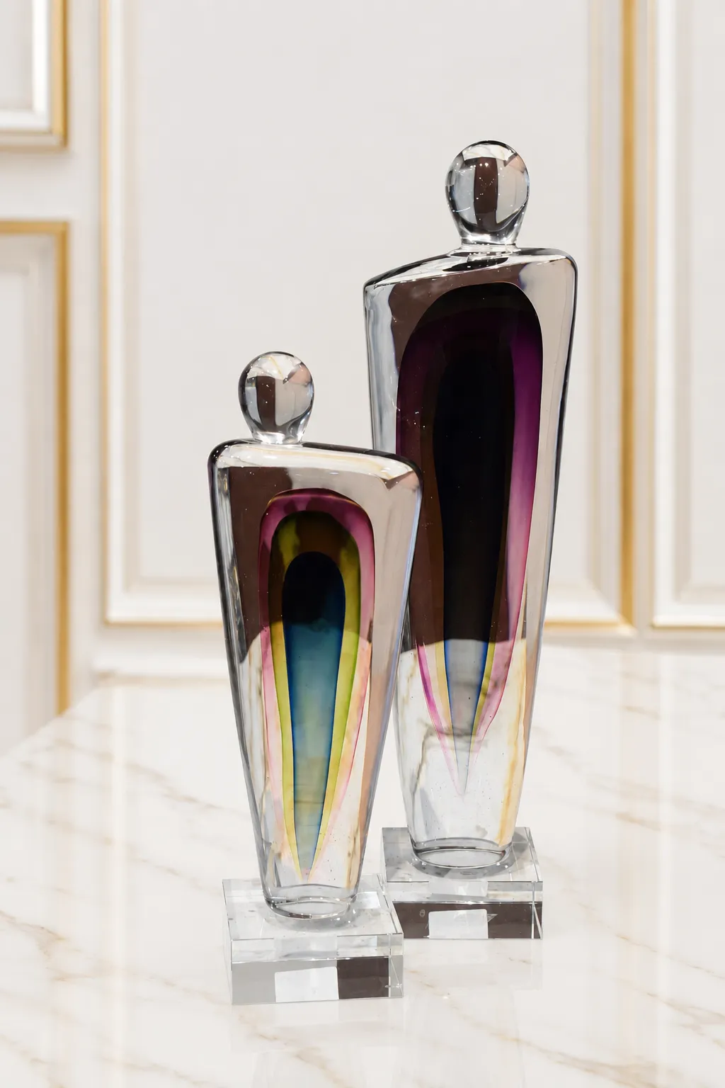 Contemporary Abstract Colored Glass Figurine Set (Pair) A pair of abstract art glass sculptures of differing heights, featuring clear thick glass shapes with embedded multi-colored cores of purple, blue, yellow, and pink, standing on square crystal bases on a marble table