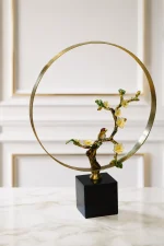 An elegant tabletop sculpture featuring an enameled cherry blossom branch with white flowers and a small bird, framed by a large golden metal ring on a black square base, placed on a light-colored marble tabletop