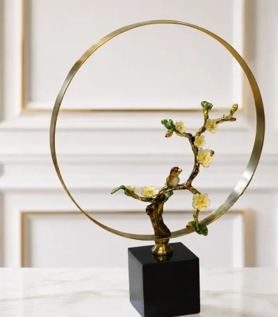 An elegant tabletop sculpture featuring an enameled cherry blossom branch with white flowers and a small bird, framed by a large golden metal ring on a black square base, placed on a light-colored marble tabletop