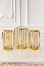 A set of three modern hurricane candle holders featuring gold wire metal cage frames with inner amber-tinted glass cylinders, arranged on a white marble table surface