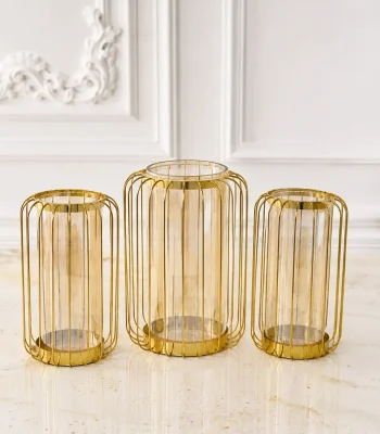 A set of three modern hurricane candle holders featuring gold wire metal cage frames with inner amber-tinted glass cylinders, arranged on a white marble table surface