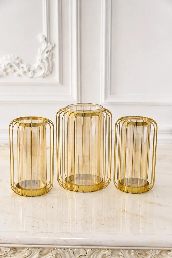 A set of three modern hurricane candle holders featuring gold wire metal cage frames with inner amber-tinted glass cylinders, arranged on a white marble table surface