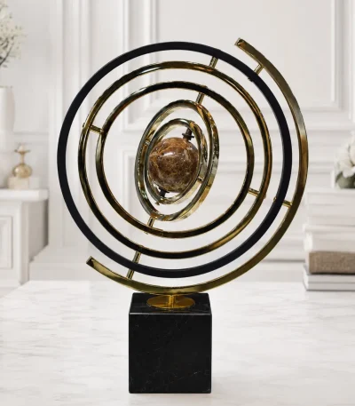 A modern kinetic-style armillary sphere sculpture featuring concentric gold and black metal rings surrounding a brown marbled central orb, standing on a black square base on a light marble tabletop