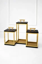 A set of three open-frame geometric lanterns made of polished gold metal bars with flat black tops and bases, each featuring a golden T-shaped handle, arranged on a light marble tabletop