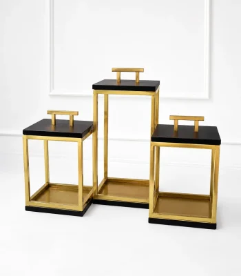 A set of three open-frame geometric lanterns made of polished gold metal bars with flat black tops and bases, each featuring a golden T-shaped handle, arranged on a light marble tabletop