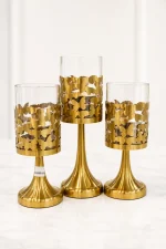 A set of three brushed gold pedestal candle holders of varying heights, featuring intricate laser-cut metal butterfly patterns wrapping around amber-tinted glass cylinder tops, standing on a light marble tabletop