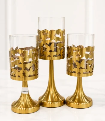 A set of three brushed gold pedestal candle holders of varying heights, featuring intricate laser-cut metal butterfly patterns wrapping around amber-tinted glass cylinder tops, standing on a light marble tabletop