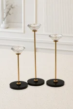 A set of three modern candlestick holders of staggered heights featuring large, clear, diamond-cut crystal cups on thin gold metal stems with circular black bases, arranged on a light-colored marble tabletop