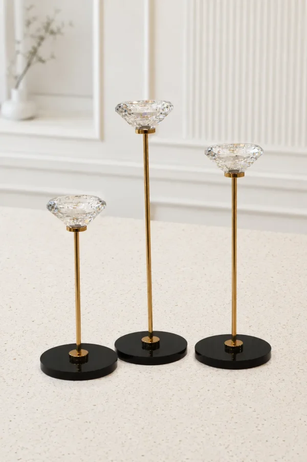 A set of three modern candlestick holders of staggered heights featuring large, clear, diamond-cut crystal cups on thin gold metal stems with circular black bases, arranged on a light-colored marble tabletop