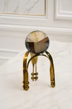 A flawless, clear crystal sphere resting on an architectural gold metal tripod stand with decorative rivet details, positioned on a light marble tabletop