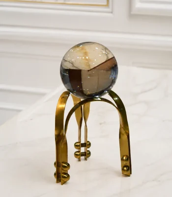 A flawless, clear crystal sphere resting on an architectural gold metal tripod stand with decorative rivet details, positioned on a light marble tabletop