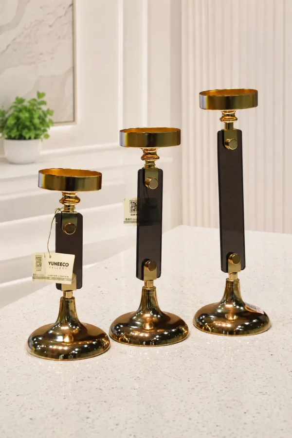 A set of three modern candlestick holders of graduated heights featuring polished gold candle cups and trumpet bases connected by flat, rectangular stems made of translucent smokey dark glass, displayed on a light marble tabletop