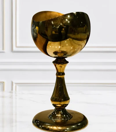 A luxury goblet-style tabletop chalice made of highly polished gold metal with a mirror-like finish and a wavy rim, standing on an ornate pedestal stem and wide circular base on a light speckled marble surface