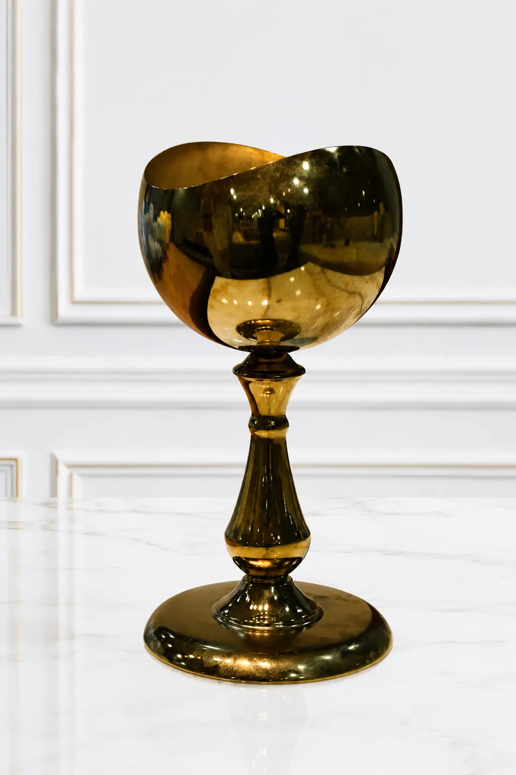 Decorative High-Shine Gold Metal Chalice A luxury goblet-style tabletop chalice made of highly polished gold metal with a mirror-like finish and a wavy rim, standing on an ornate pedestal stem and wide circular base on a light speckled marble surface