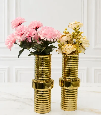 A matching pair of cylindrical metallic gold ceramic vases featuring horizontal ribbed textures and a smooth wider center ring, holding pink and white faux flowers on a light marble tabletop