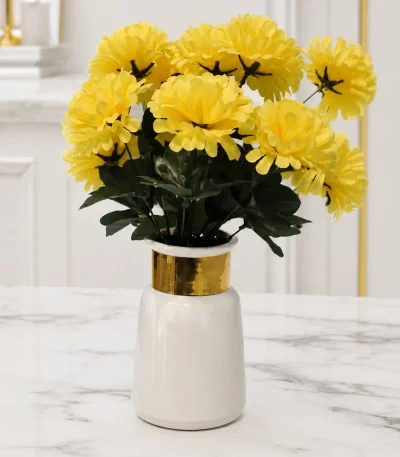 A modern white ceramic vase with a glossy finish and a thick polished gold band around its neck, holding a bouquet of faux yellow flowers on a light marble table