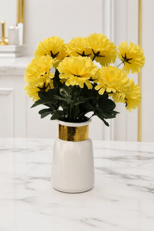 A modern white ceramic vase with a glossy finish and a thick polished gold band around its neck, holding a bouquet of faux yellow flowers on a light marble table