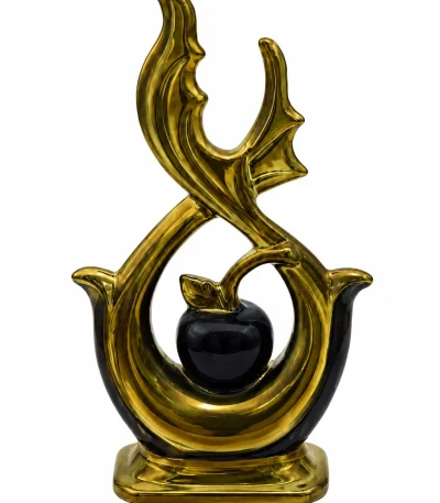 An elegant abstract ceramic sculpture featuring high-gloss gold metallic flame-like loops framing a central black apple with a leaf, standing on a round wood-grain accent pedestal table