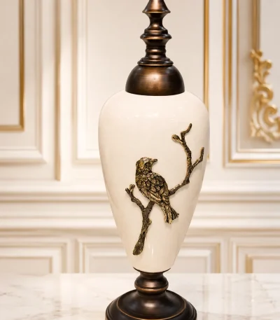 Luxury white ceramic decorative jar with a bronze-toned lid and an embossed bird perched on a branch, displayed on a marble tabletop.