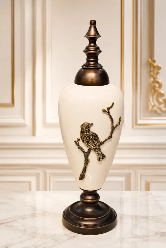 Luxury white ceramic decorative jar with a bronze-toned lid and an embossed bird perched on a branch, displayed on a marble tabletop.