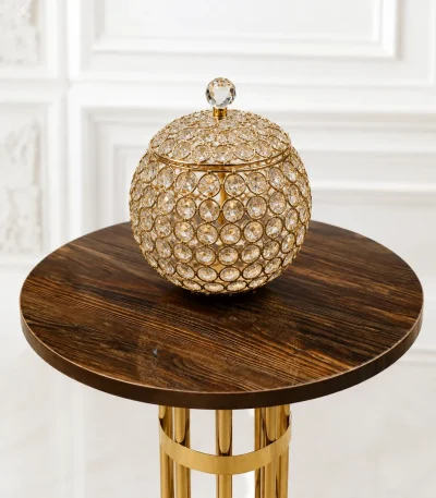 A luxury spherical decorative jar crafted from a gold-plated metal wire lattice woven with multifaceted clear crystal beads, complete with a matching lidded top and crystal finial knob, sitting on a round wood-grain pedestal table