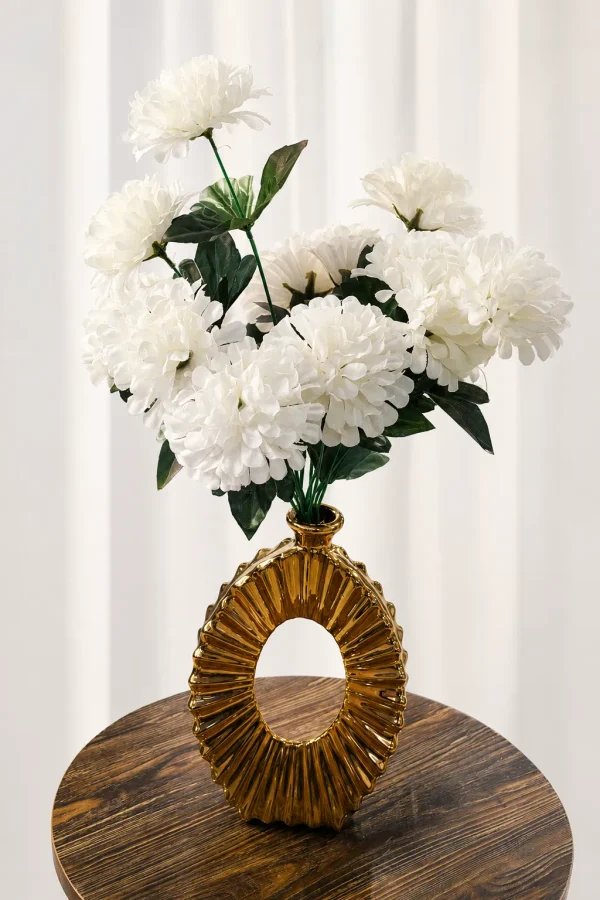 A contemporary metallic gold ceramic vase with a hollow oval doughnut shape and a deep, radiating ribbed texture, holding a full bouquet of white faux flowers on a round dark wood side table