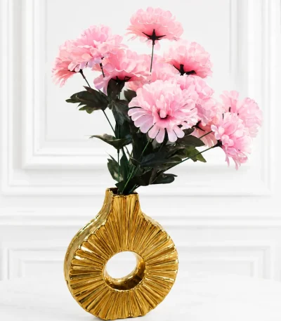 A contemporary metallic gold ceramic donut vase featuring a hollow circular shape and a radiating ribbed texture, holding a bouquet of fluffy faux pink flowers on a round wooden side table