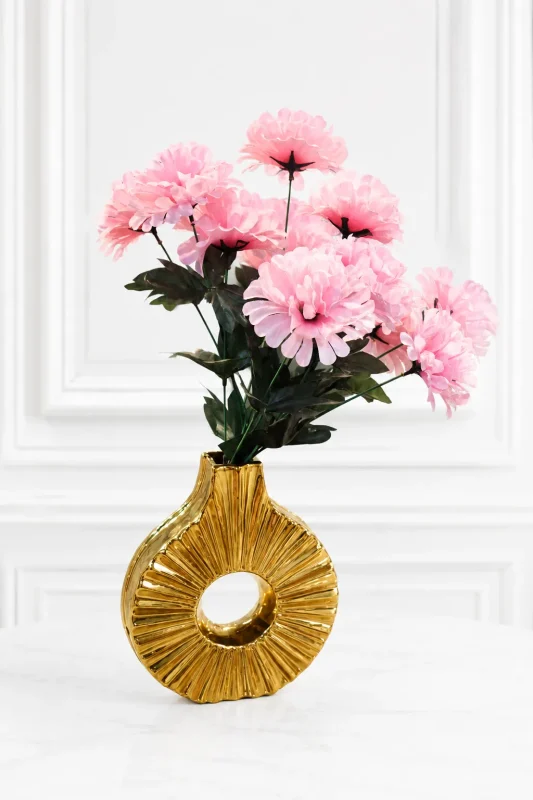 A contemporary metallic gold ceramic donut vase featuring a hollow circular shape and a radiating ribbed texture, holding a bouquet of fluffy faux pink flowers on a round wooden side table