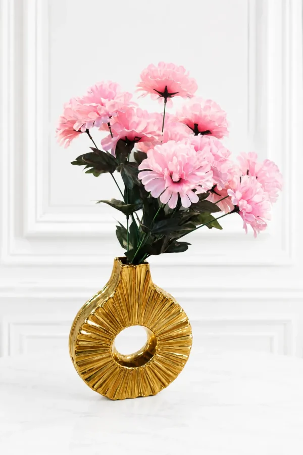 A contemporary metallic gold ceramic donut vase featuring a hollow circular shape and a radiating ribbed texture, holding a bouquet of fluffy faux pink flowers on a round wooden side table