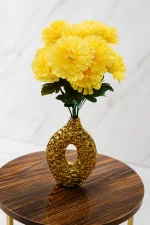 A contemporary metallic gold ceramic vase with a hollow oval doughnut shape and a heavily dimpled, hammered texture, holding a full bouquet of bright yellow faux flowers on a round wooden table