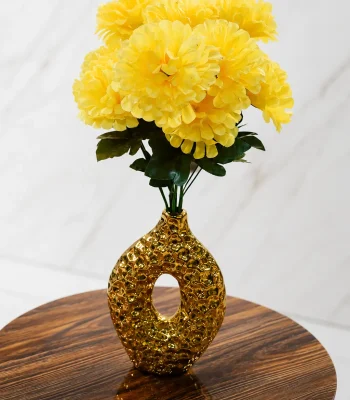 A contemporary metallic gold ceramic vase with a hollow oval doughnut shape and a heavily dimpled, hammered texture, holding a full bouquet of bright yellow faux flowers on a round wooden table