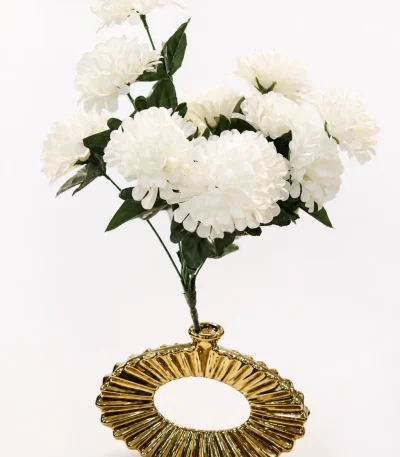 A contemporary metallic gold ceramic vase with a hollow oval doughnut shape and a deep, radiating ribbed texture, holding a full bouquet of white faux flowers on a round dark wood side table