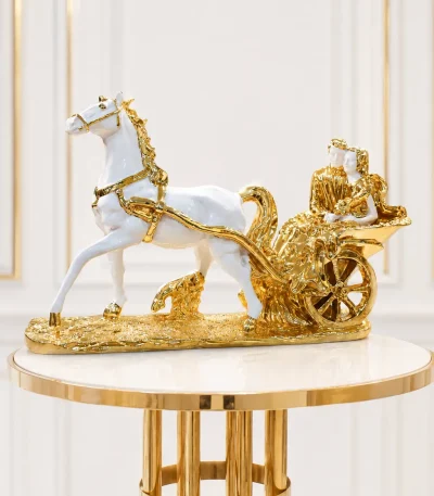 An ornate luxury decorative statue featuring a glossy white horse with gold hooves and harness pulling an elaborate metallic gold carriage containing a couple, all secured to a textured golden pedestal base on a round wood-grain table