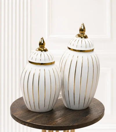 A set of two decorative white ceramic ginger jars of varying sizes, featuring vertical gold stripes and polished gold finial lids, sitting together on a round wood-grain table