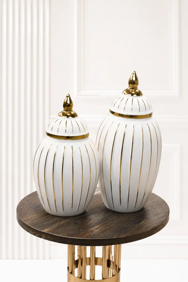 A set of two decorative white ceramic ginger jars of varying sizes, featuring vertical gold stripes and polished gold finial lids, sitting together on a round wood-grain table
