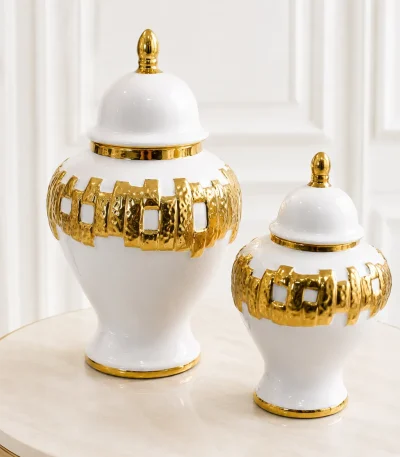 A set of two decorative white ceramic ginger jars of varying sizes, featuring vertical gold stripes and polished gold finial lids, sitting together on a round wood-grain table