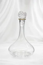 An elegant clear glass wine decanter with a wide, rounded base and a long narrow neck accented by a gold rim ring, topped with a large, ornate diamond-cut faceted crystal stopper, displayed on a beige marble table