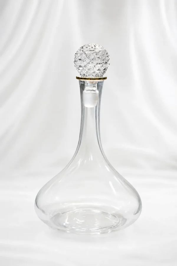 An elegant clear glass wine decanter with a wide, rounded base and a long narrow neck accented by a gold rim ring, topped with a large, ornate diamond-cut faceted crystal stopper, displayed on a beige marble table