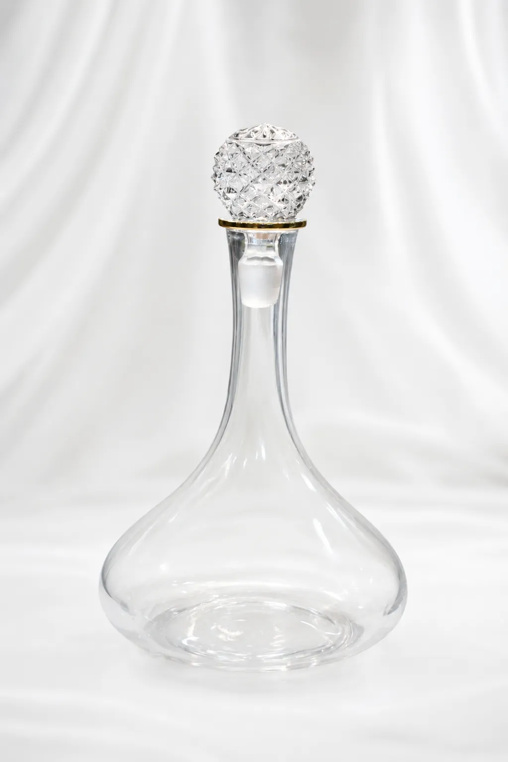 Luxury Spirits and Wine Carafe An elegant clear glass wine decanter with a wide, rounded base and a long narrow neck accented by a gold rim ring, topped with a large, ornate diamond-cut faceted crystal stopper, displayed on a beige marble table