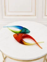 A pair of decorative, hand-blown art glass fish figurines featuring stylized curved bodies and textured ridges—one shaded in blue and green, and the other in red and amber yellow—displayed together on a round beige marble side table