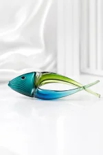 A decorative, hand-blown art glass fish figurine featuring a stylized curved body split into deep blue and translucent green, with fine textured ridges on its teal head, displayed on a beige marble table