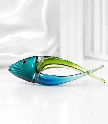 A decorative, hand-blown art glass fish figurine featuring a stylized curved body split into deep blue and translucent green, with fine textured ridges on its teal head, displayed on a beige marble table