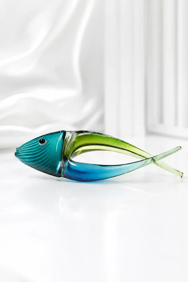 A decorative, hand-blown art glass fish figurine featuring a stylized curved body split into deep blue and translucent green, with fine textured ridges on its teal head, displayed on a beige marble table