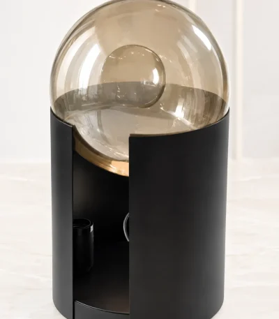 A contemporary decorative table lamp featuring a large, transparent glass sphere dome nestled inside a curved matte black cylindrical metal base with a hidden gold accent ring, resting on a beige marble surface