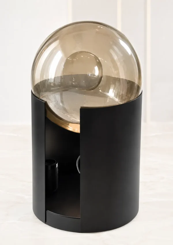 A contemporary decorative table lamp featuring a large, transparent glass sphere dome nestled inside a curved matte black cylindrical metal base with a hidden gold accent ring, resting on a beige marble surface