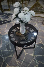 Round black metal coffee table with a tempered glass top and decorative gear design, displayed with a white flower vase.