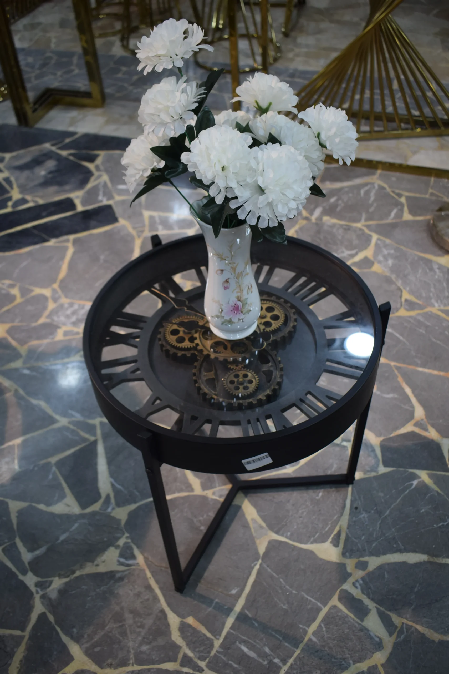Modern Black Metal Gear Coffee Table | Round Glass Top Side Table for Living Room Round black metal coffee table with a tempered glass top and decorative gear design, displayed with a white flower vase.